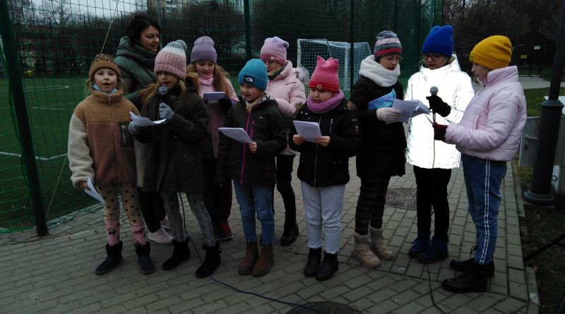 Na chodniku grupa śpiewających dzieci ubranych w ciepłe kurtki i czapki, z kartkami w rękach i z dwoma mikrofonami.
