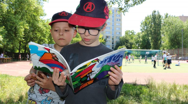 Na boisku szkolnym dwóch chłopców w czapkach z daszkiem czyta komiks. W tle dzieci grające w piłkę nożną.