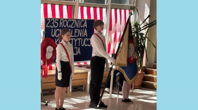 Na tle granatowej tablicy z białym napisem biało - czerwonej dekoracji uczeń ze sztandarem i dwie uczennice w strojach galowych.